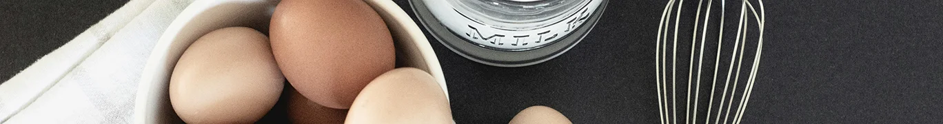 Brown eggs in a white bowl next to a glass pitcher of milk on a dark surface.