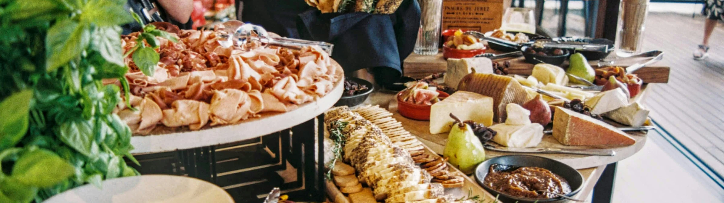 A buffet table with assorted meats, cheeses, breads, and fruits, with people conversing in the background.
