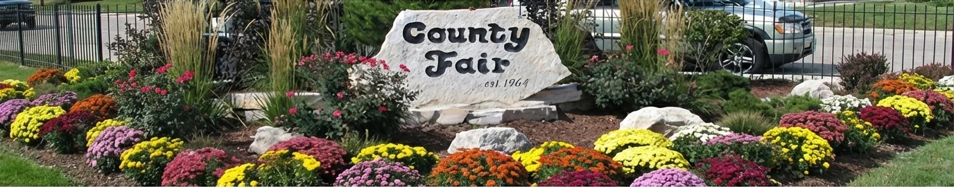 Stone sign in a flower garden reads County Fair est. 1964 with colorful flowers around it.